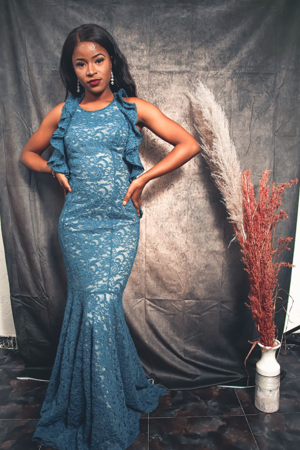 Long blue lace evening dress with a ruffled neckline and drop earrings against a gray studio backdrop