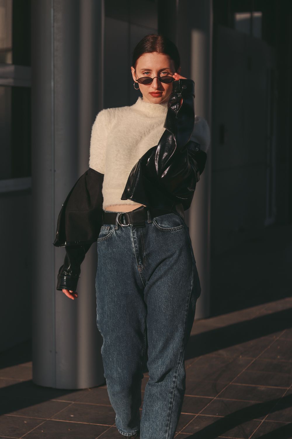 Black jacket over a cream knit top with relaxed blue jeans and sunglasses on a city street
