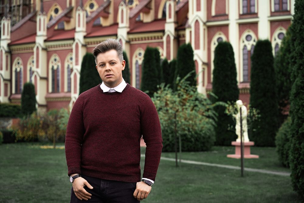 Layering detail of a burgundy sweater over a white collared shirt with dark trousers outdoors